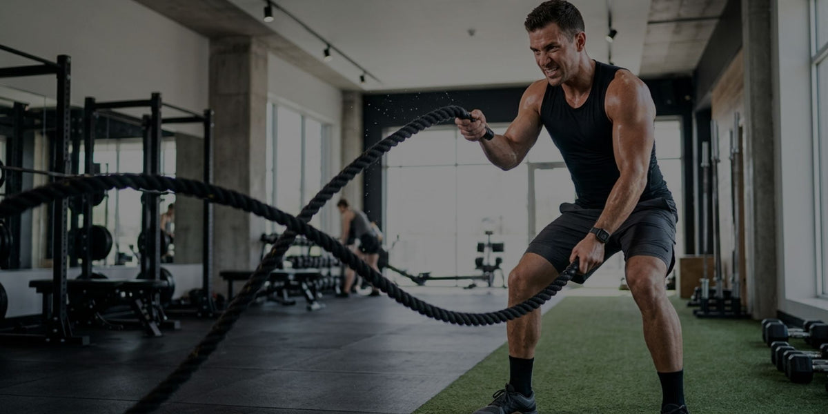 A man in a gym wearing a black tank top and shorts, performing a battle rope exercise with a thick black battle rope. The gym features dark flooring, turf, and various exercise equipment in the background.